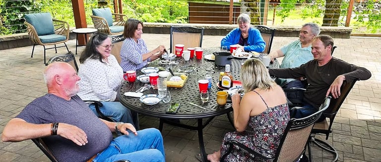 Group of eight people seated around a round patio table enjoying a meal and conversation outdoors