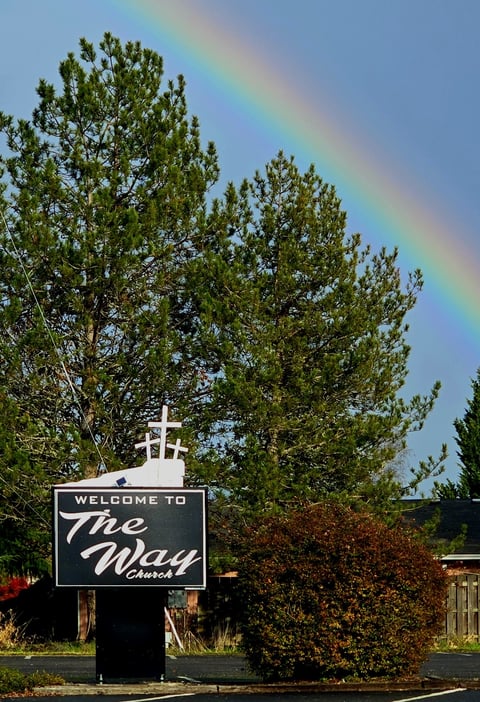 Welcome to The Way church sign with cross beneath tall pine trees and rainbow in blue sky